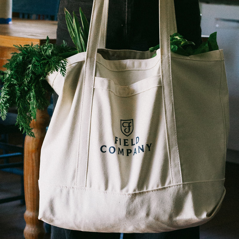 Canvas Field Market Tote with navy shield logo, loaded with carrot tops and leafy greens, carried indoors