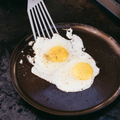 Two sunny-side-up eggs cooking on the No.9 Field Round Griddle, slotted turner easing under one thumbnail