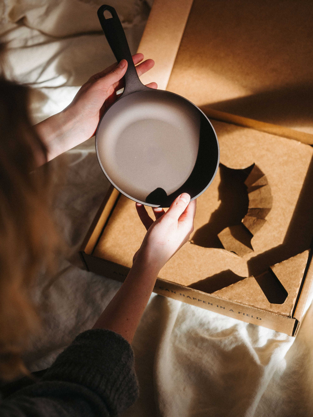 Person lifting the No.5 Field Cast Iron Skillet from its kraft box while seated on a bed in morning light