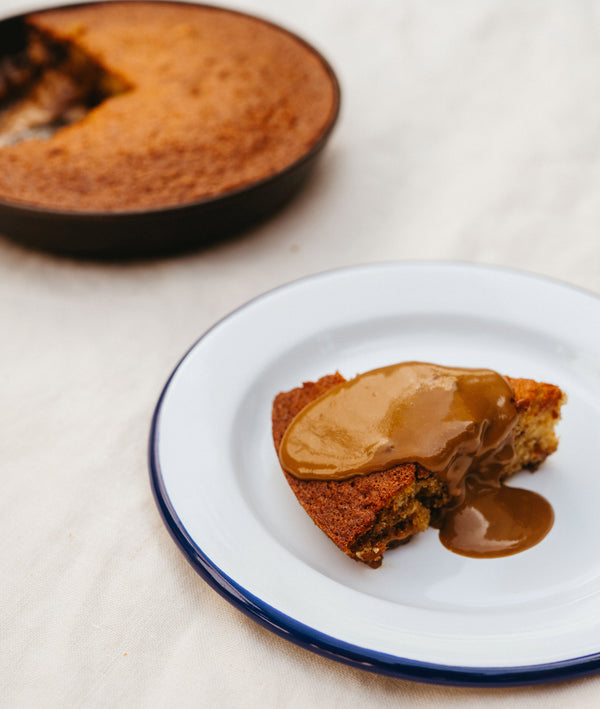 Slice of cake smothered in warm toffee sauce on an enamel plate, the rest of the cake in a Field Skillet behind