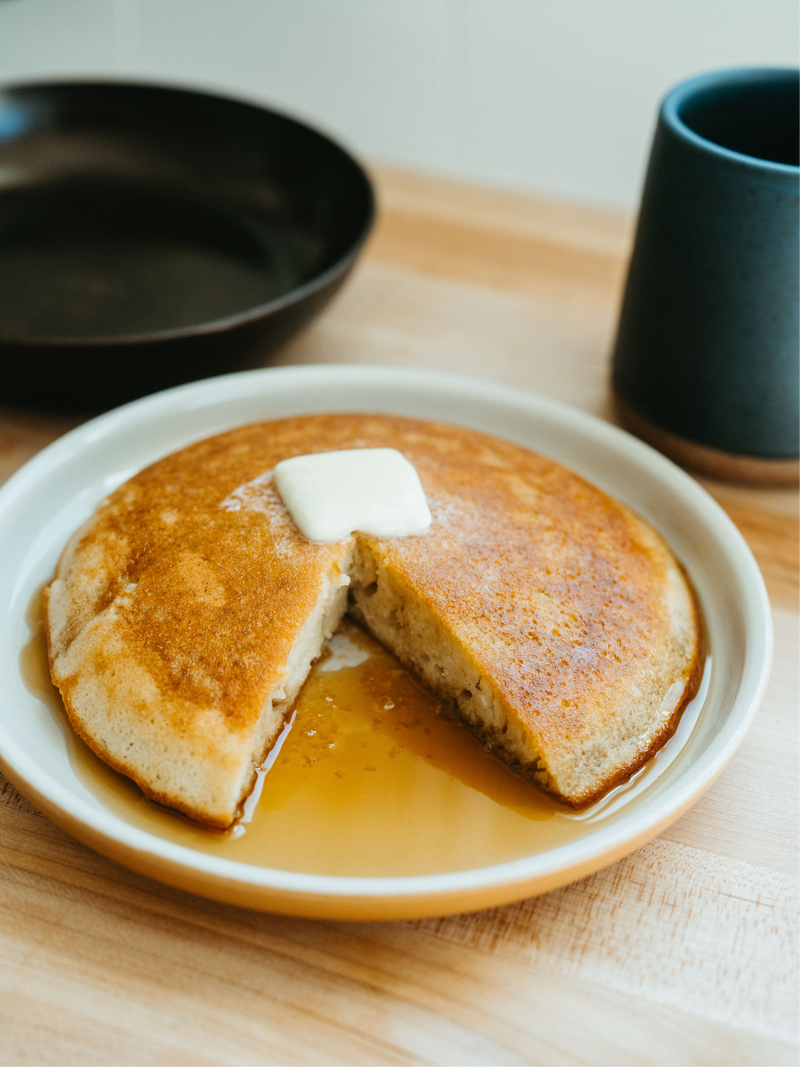 Thick skillet pancake topped with butter and syrup on a plate, one wedge cut, with a mug and skillet behind