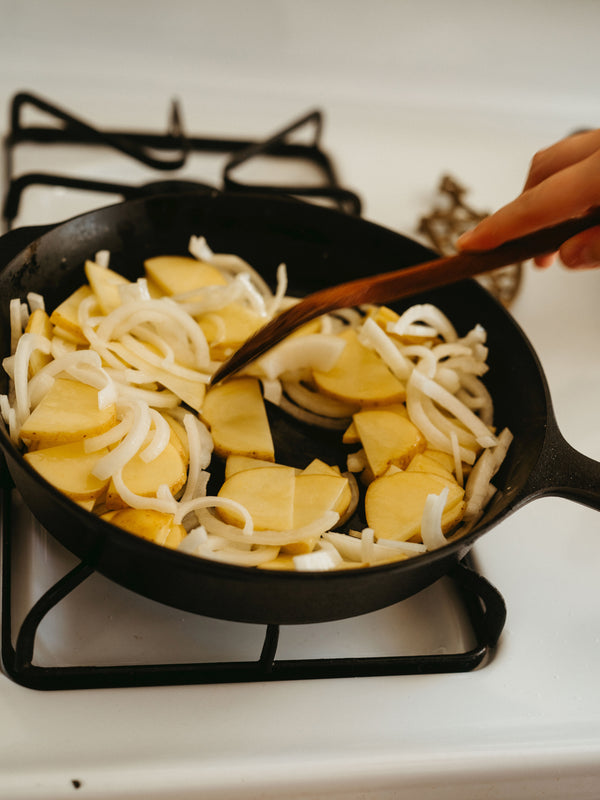 Person stirring sliced potatoes and onions in a No.5 Field Cast Iron Skillet with a wooden spatula