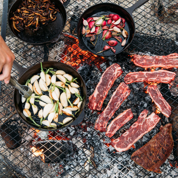 Field Company cast iron skillets cooking vegetables over an open fire next to grilling short ribs on a campfire grate