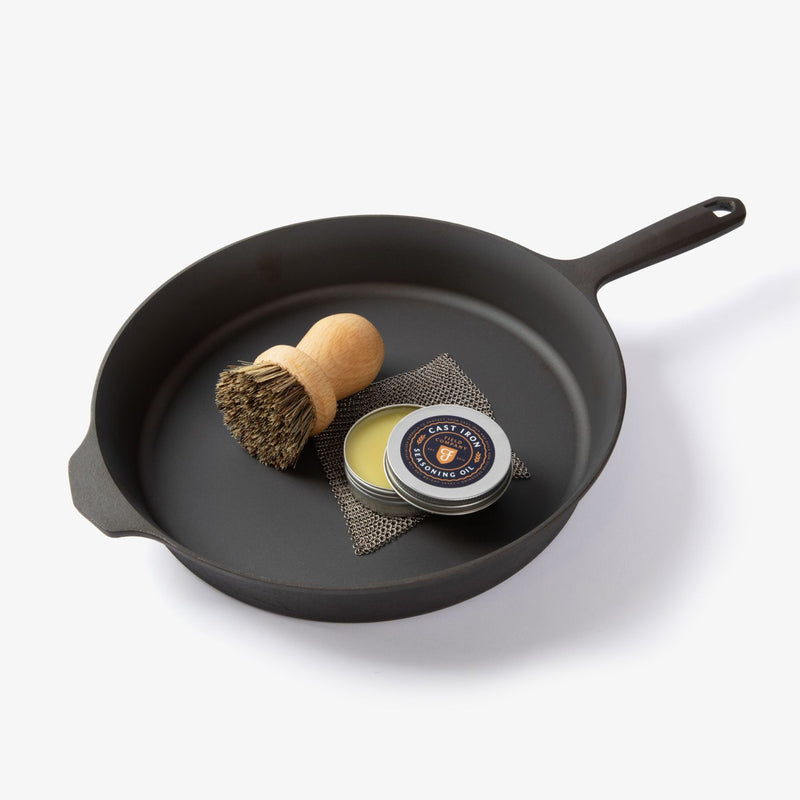Natural fiber brush, chain mail scrubber, and open tin of Field Company Cast Iron Seasoning Oil inside a cast iron skillet