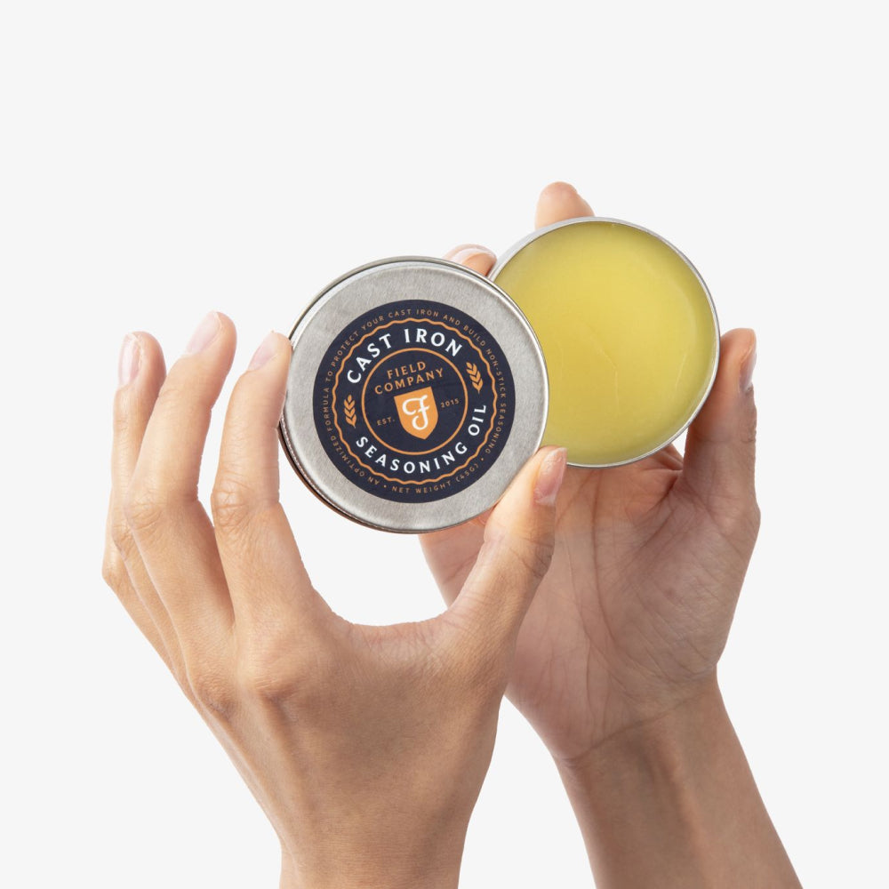 Hands holding an open tin of Field Company Cast Iron Seasoning Oil, showing the labeled lid and balm inside