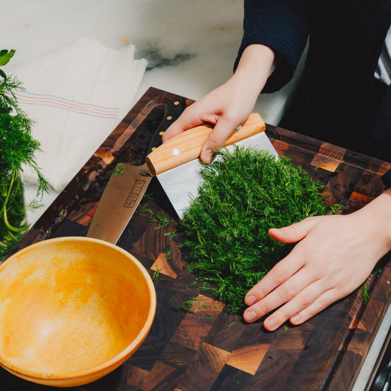 Hands scooping chopped dill with the Field Bench Scraper on an end-grain cutting board