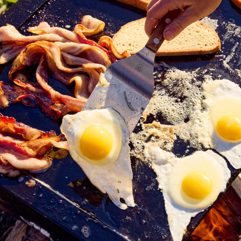 Hand flips a sunny-side-up egg with the Field Griddle Turner beside bacon and bread on an outdoor griddle