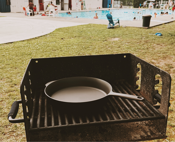 Field Skillet resting on a charcoal park grill with a swimming pool in the background