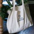 Field Market Tote filled with fresh greens beside a blue chair on a wood floor, shield logo facing out thumbnail