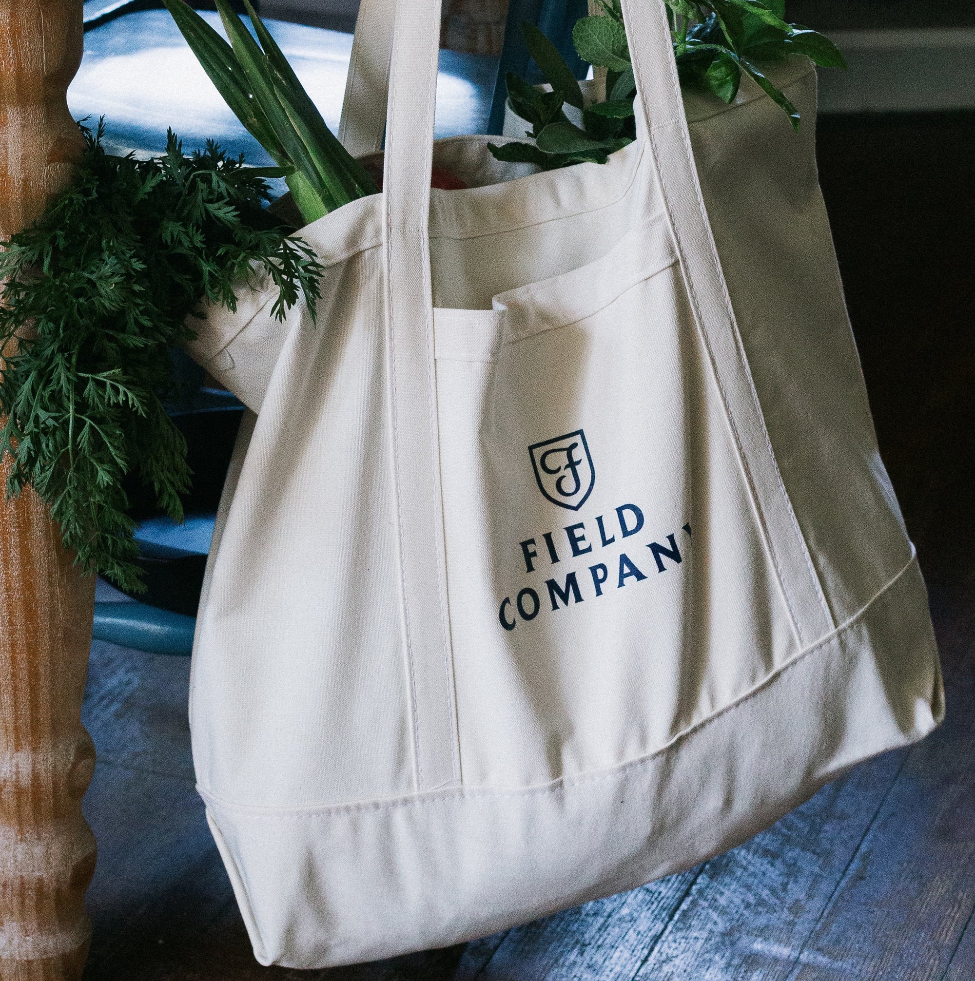 Field Market Tote filled with fresh greens beside a blue chair on a wood floor, shield logo facing out