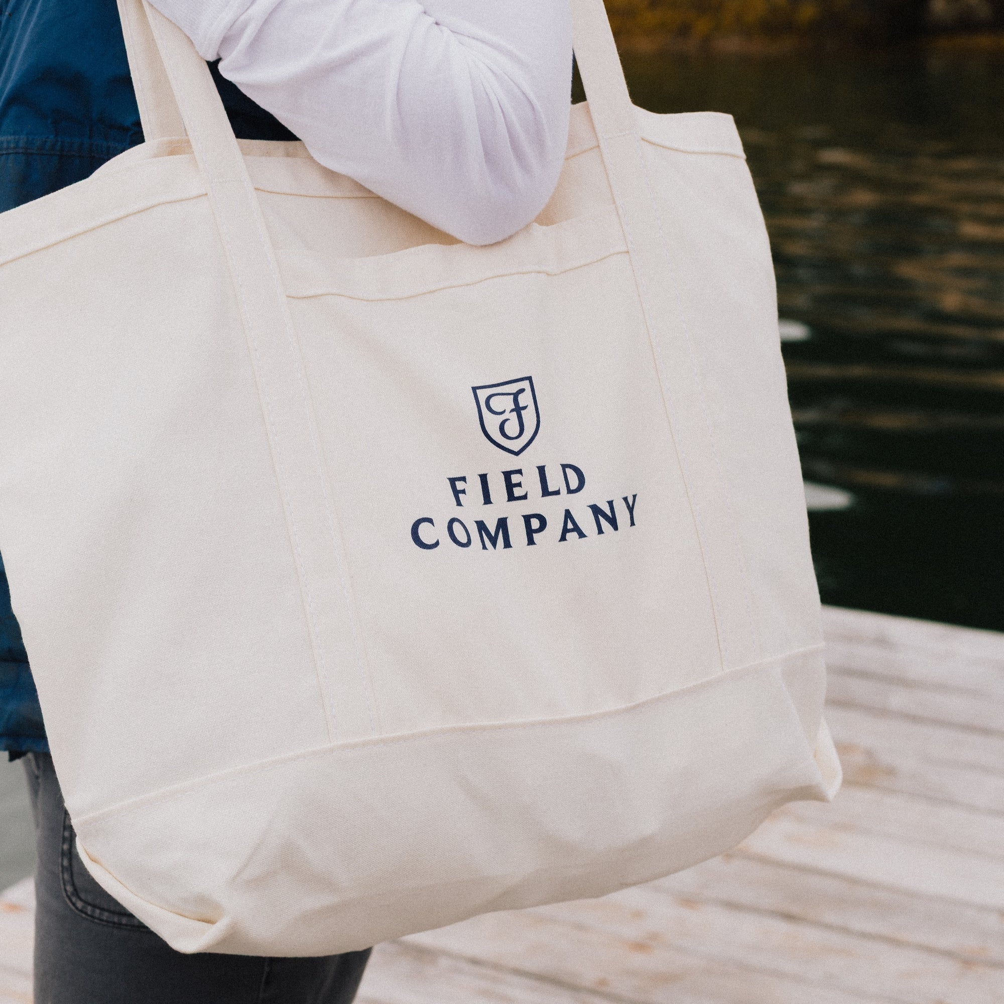 Field Market Tote in natural canvas with navy logo, carried over the shoulder on a waterside dock