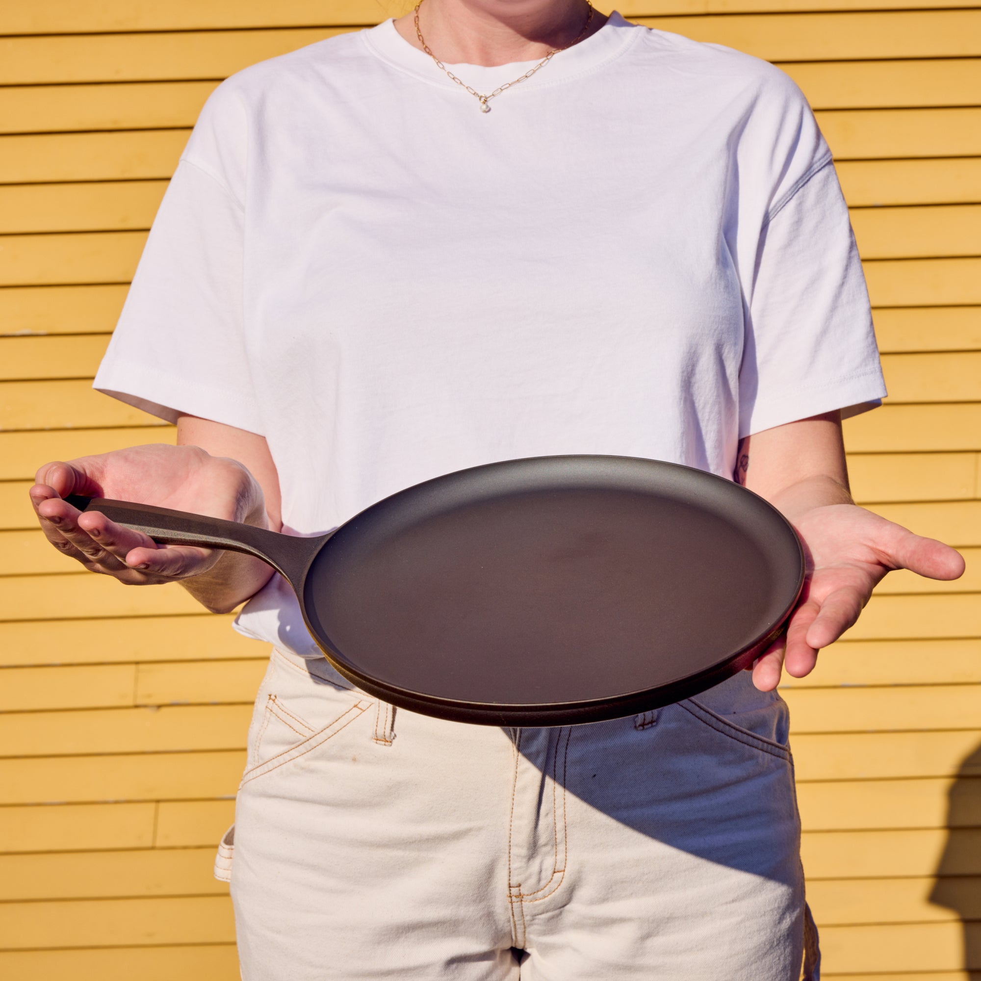 Person holding the No.9 Field Round Cast Iron Griddle flat across both hands in front of a yellow clapboard wall