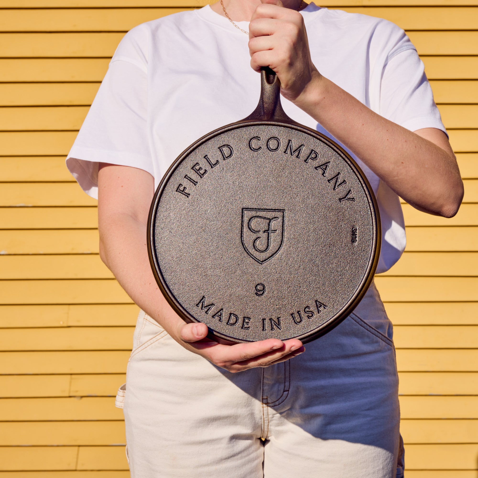 Person holding the No.9 Field Cast Iron Griddle to show its stamped underside, against yellow siding