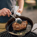 Person spoons pan juices over a ribeye steak searing in a Field Cast Iron Skillet on an outdoor grill thumbnail