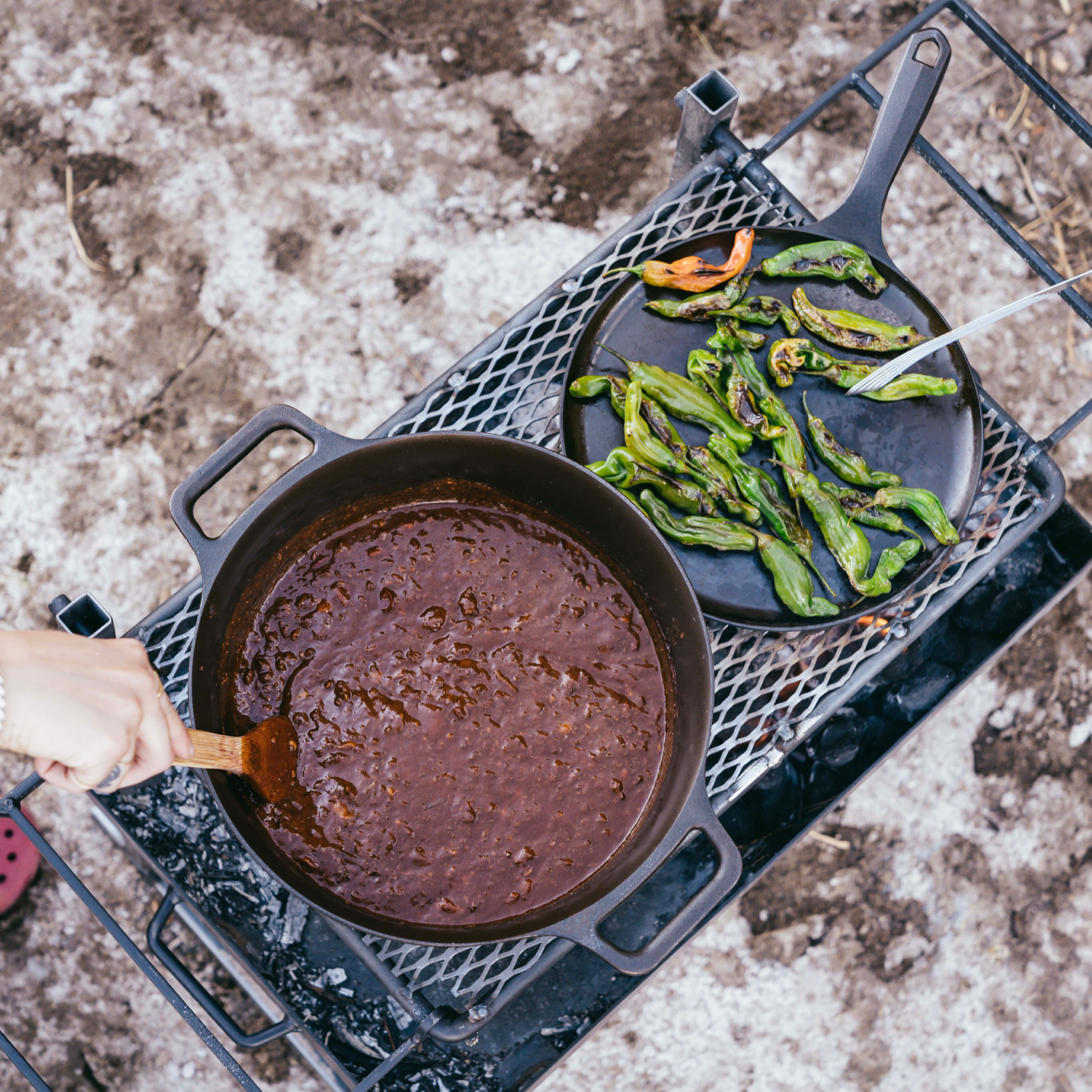 Chili simmering in the No.10 Field Dutch Oven on a campfire grate beside a plate of charred peppers