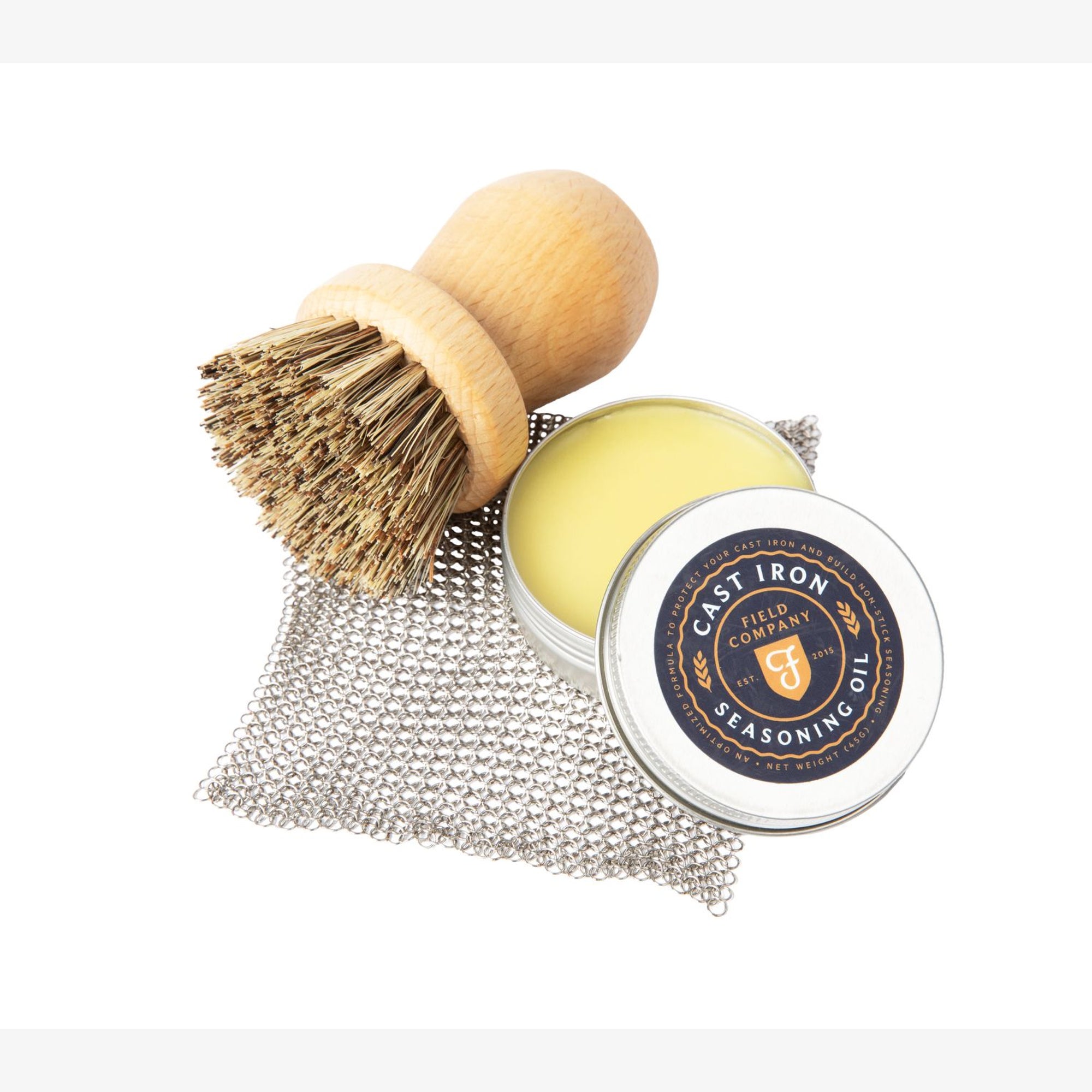 Field cast iron care tools: beech pot brush, open seasoning oil tin, and chain mail scrubber on white