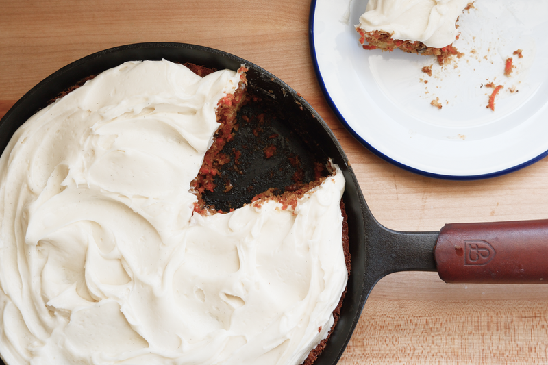 Frosted carrot cake in a Field Skillet with a red handle cover, one slice served on an enamel plate