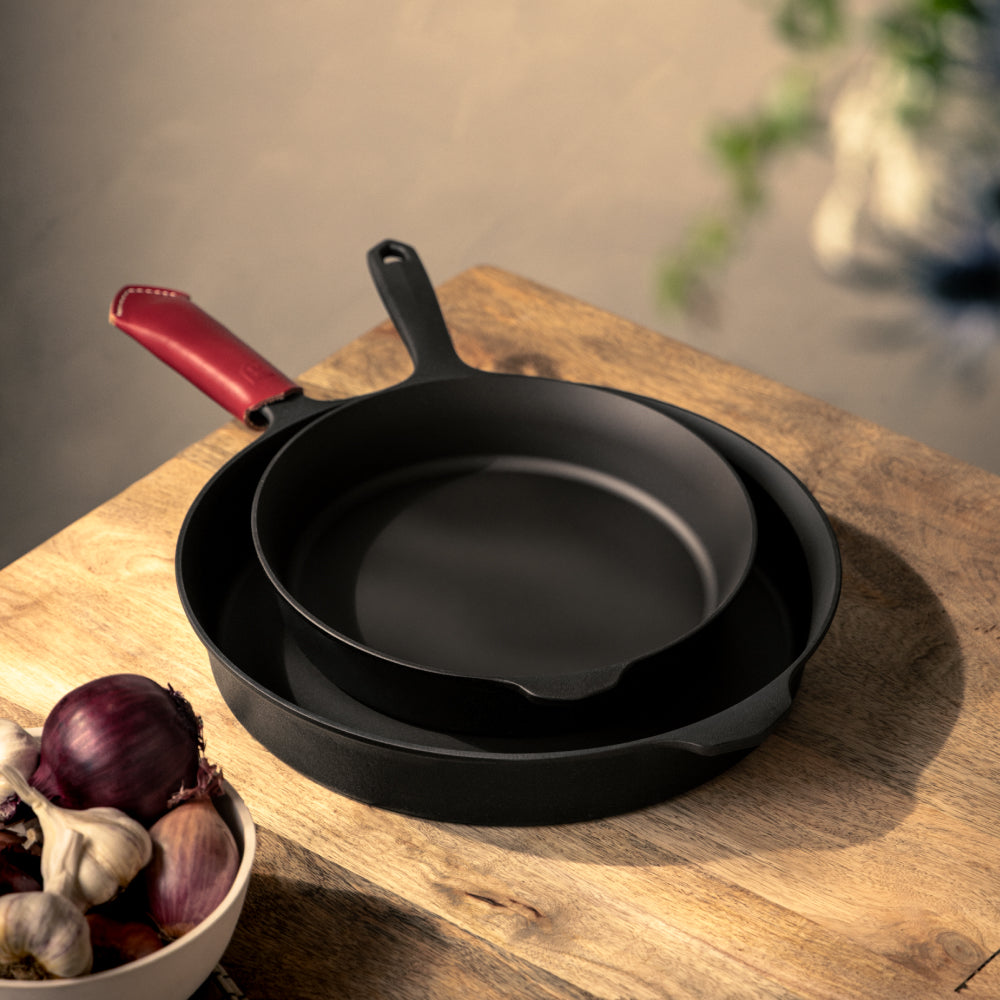 Two nested Field Skillets with a red leather handle cover on a wood board beside a bowl of onions and garlic