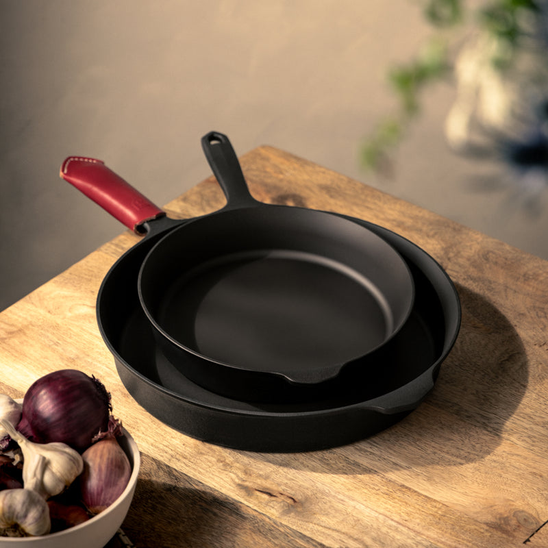 Two nested Field Skillets with a red leather handle cover on a wood board beside a bowl of onions and garlic