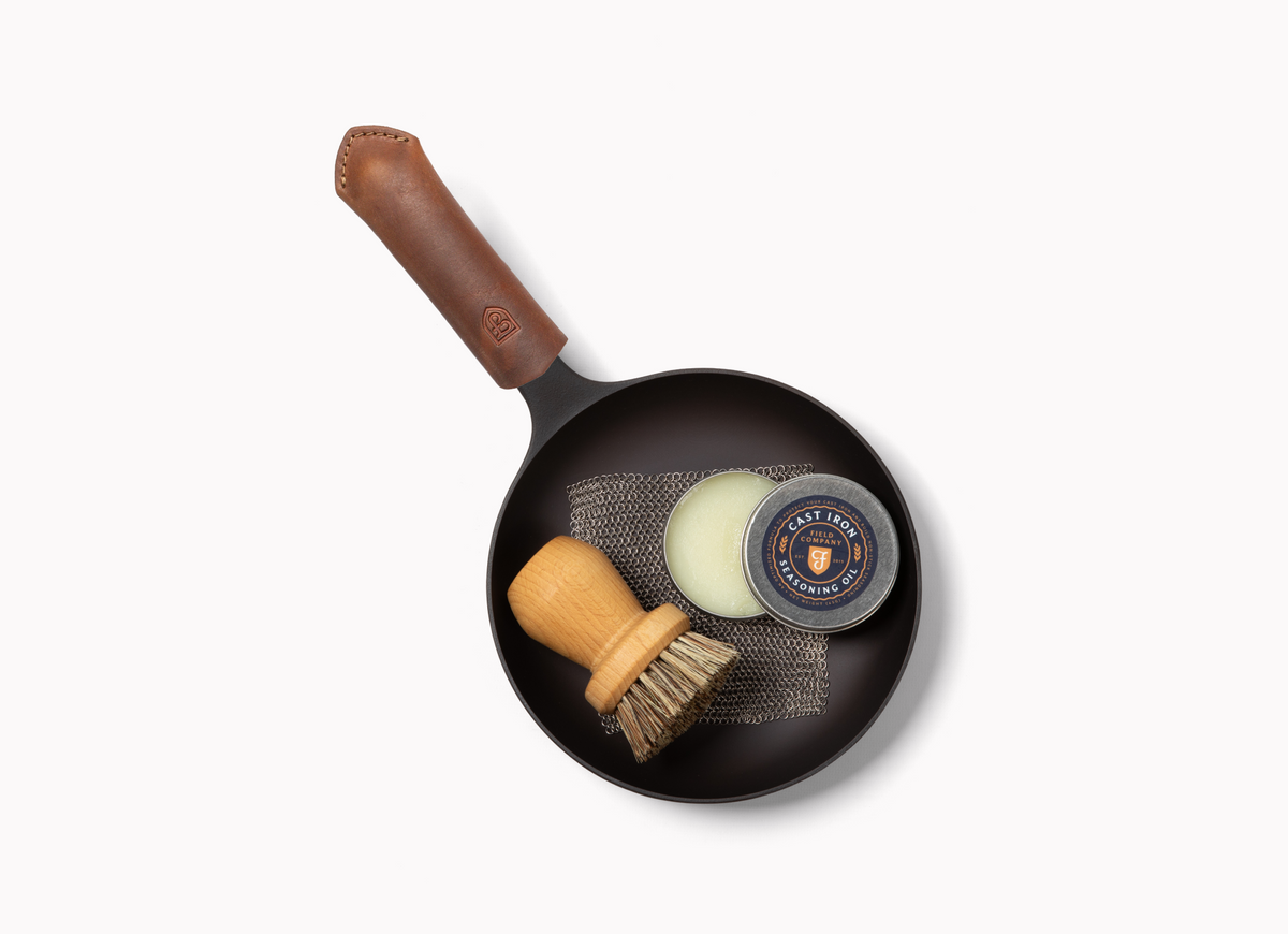 Overhead of a small Field Skillet with leather handle cover holding a scrub brush, chainmail, and seasoning oil tin