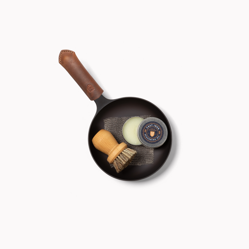 Overhead view of the Field egg pan with leather handle cover holding a scrub brush, chain mail, and seasoning oil tin