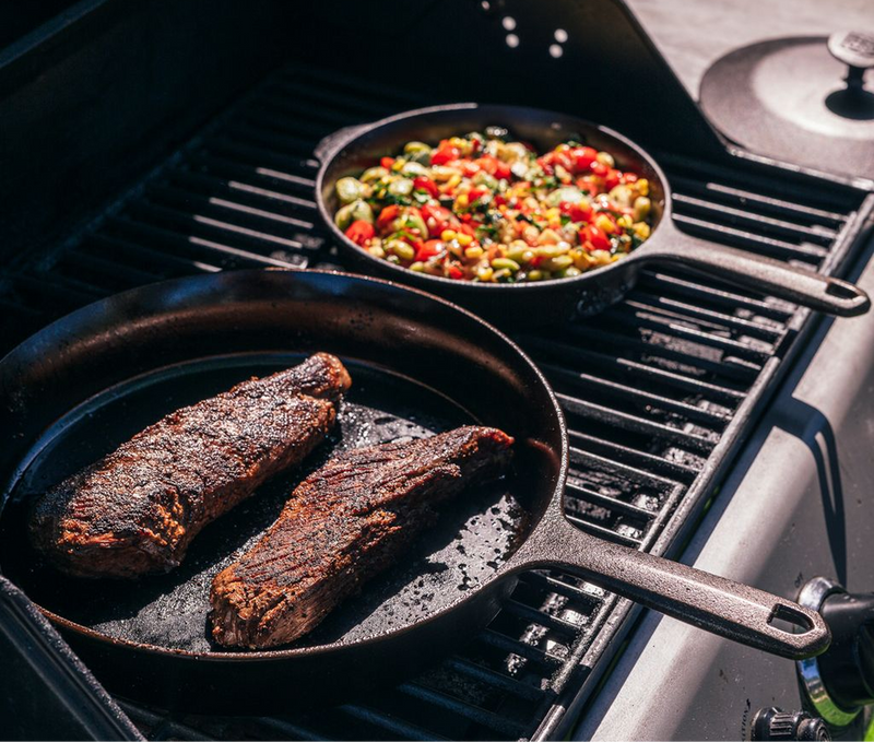 Two steaks searing in a Field Skillet on a grill beside a pan of vegetables