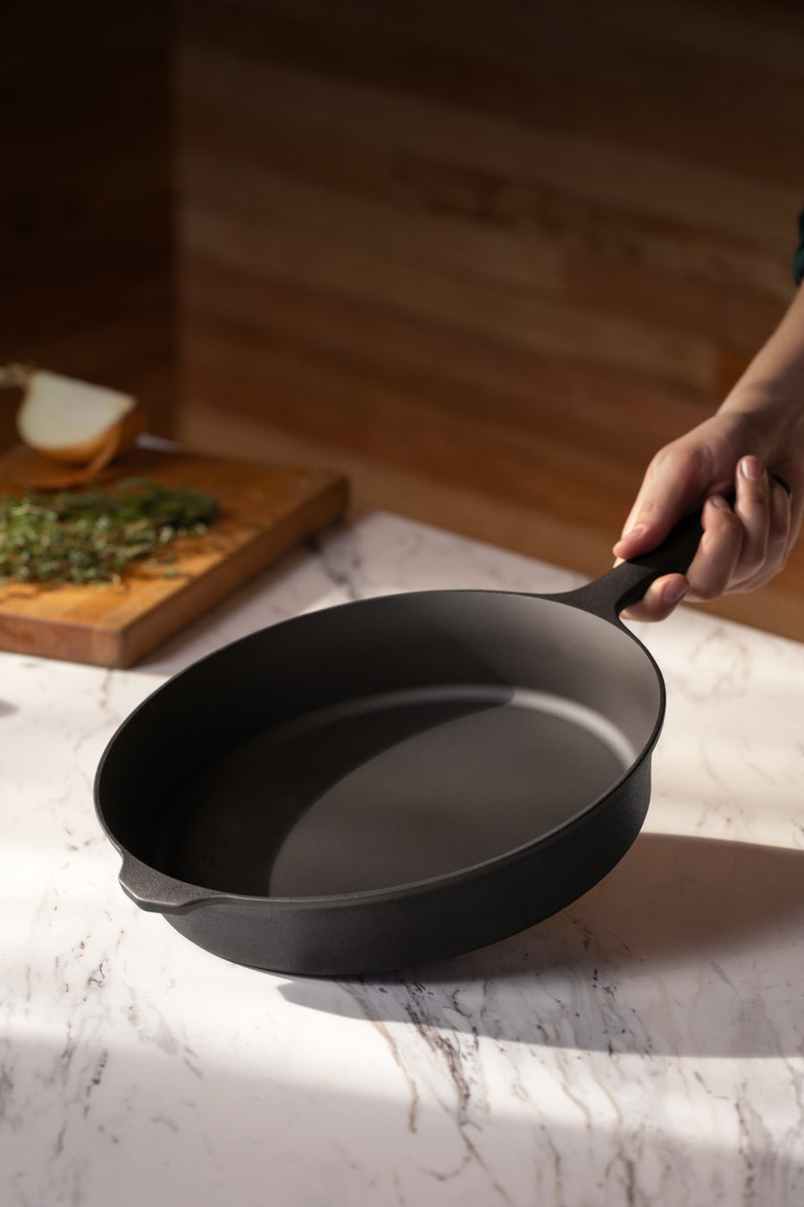 Hand lifting the No.8 Field Cast Iron Skillet from a marble counter, chopped herbs on a board behind