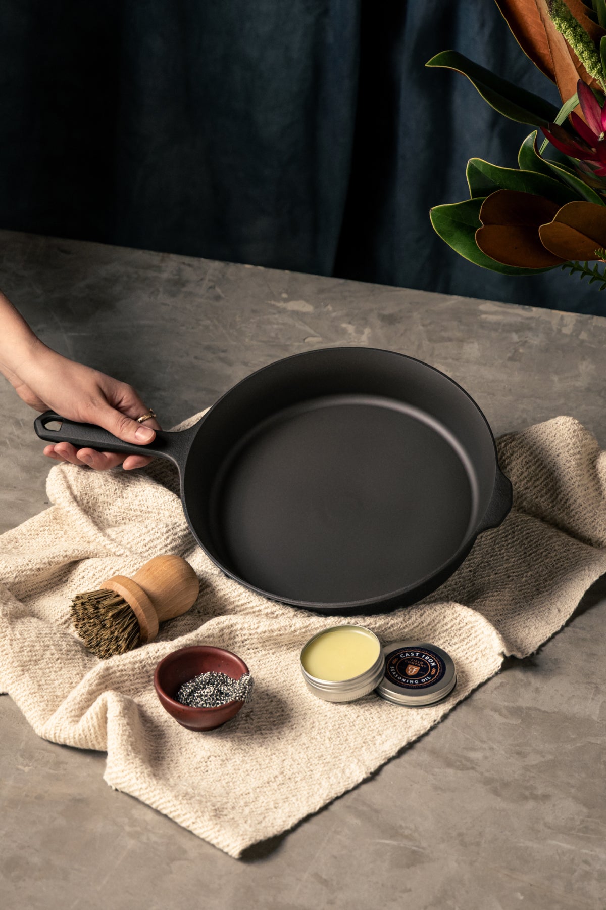 Hand holding the Field Skillet on a woven towel with a scrub brush, chainmail, and open tin of seasoning oil