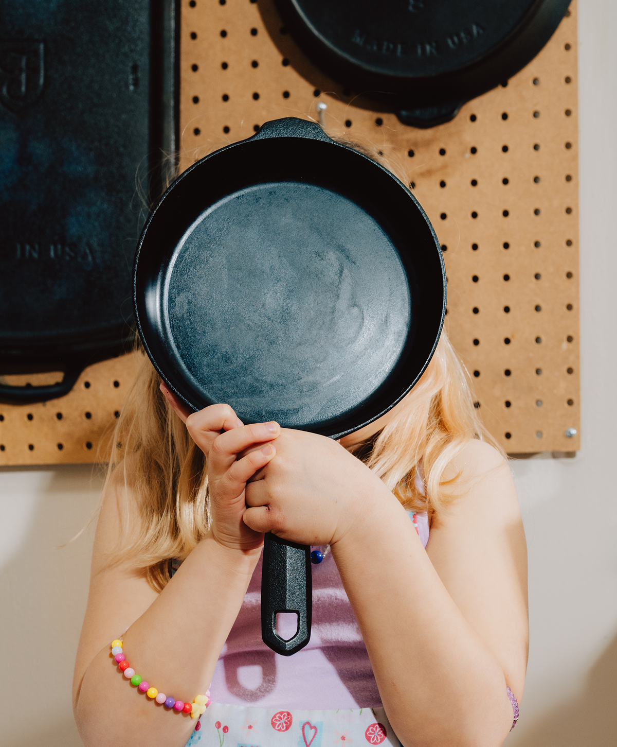 Child holding a Field Skillet in front of their face, pegboard of pans behind