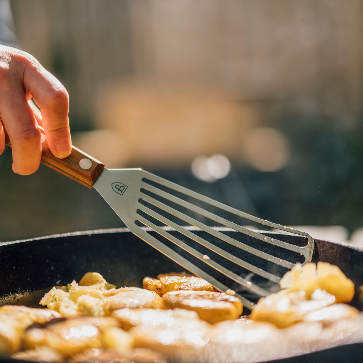 Hand flipping crispy potatoes in a cast iron pan outdoors with the Field Slotted Turner