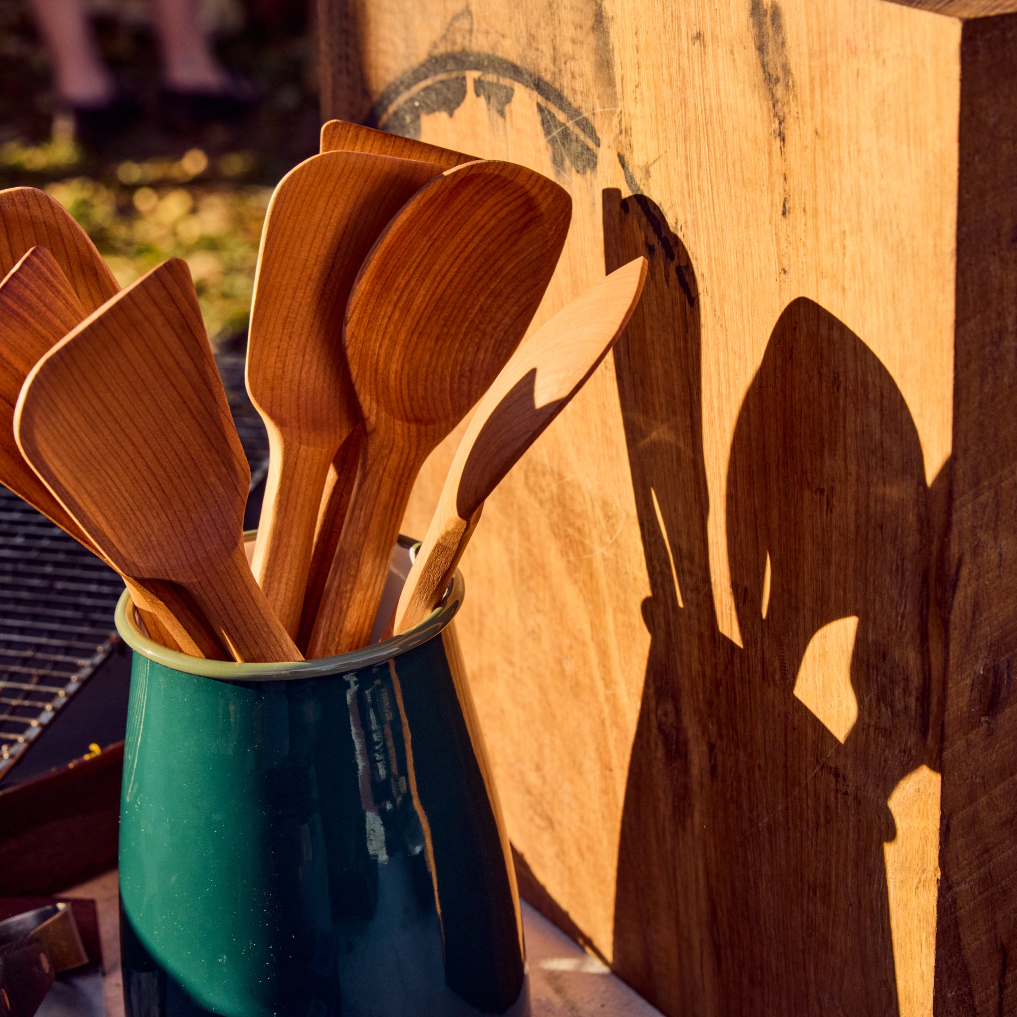 Cherry wood Field spoons and spatulas in a green enamel pitcher outdoors, casting long shadows in warm light
