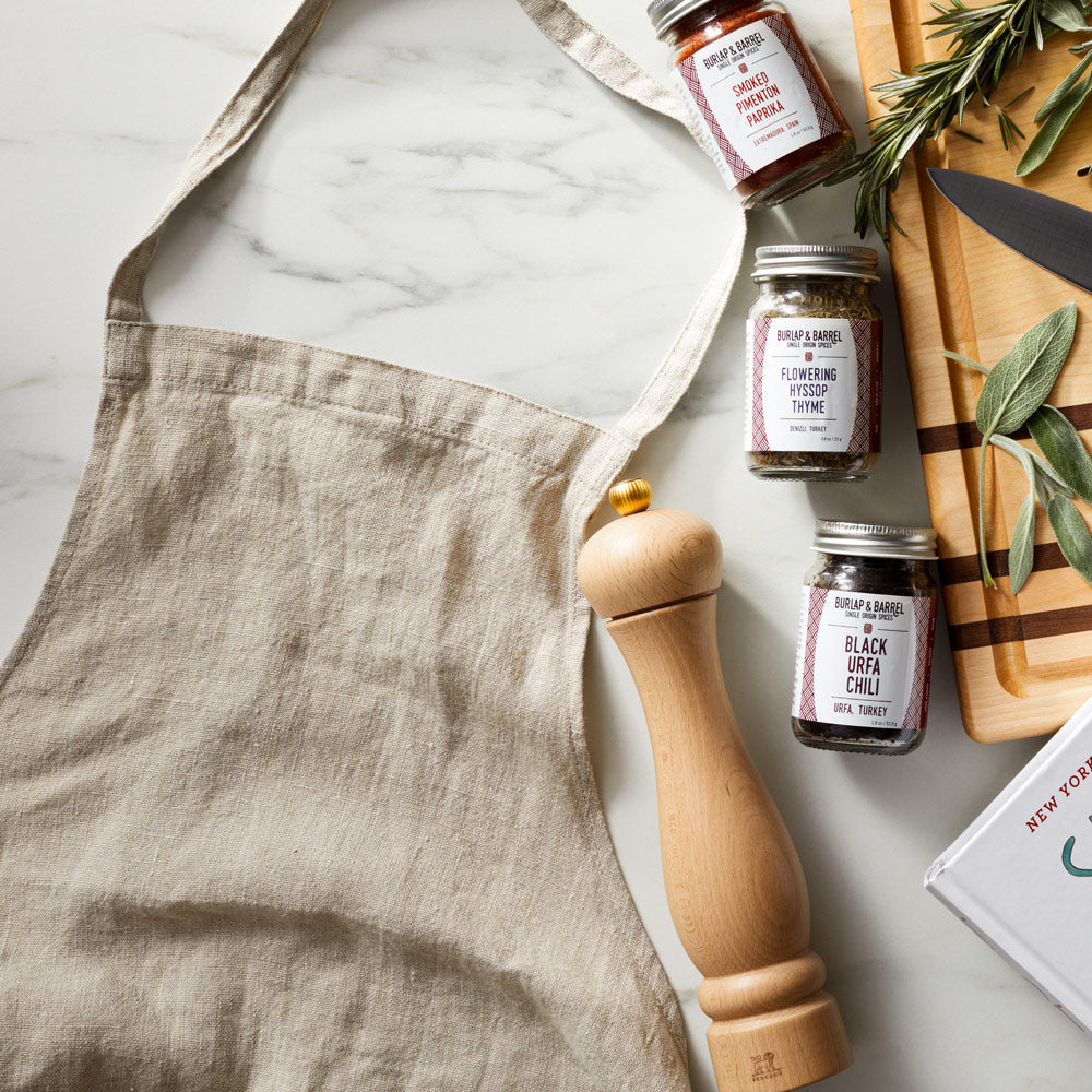 Overhead flat lay of the Field linen apron on marble with a wooden pepper mill, spice jars, and fresh herbs